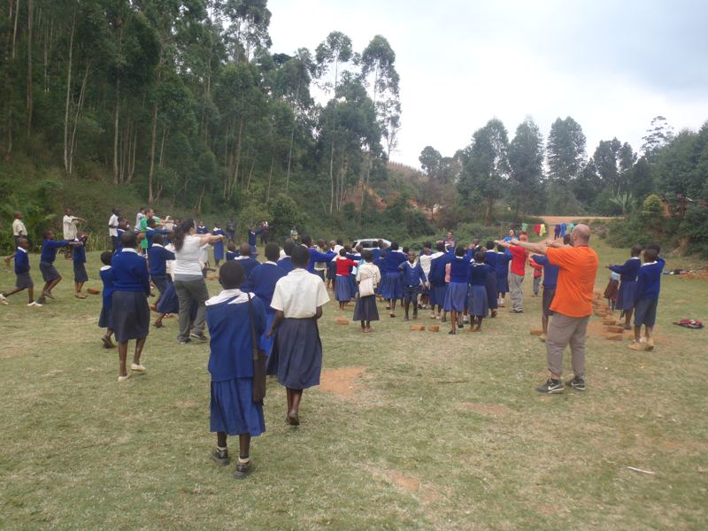 Pupils in blue school uniforms play a game with volunteers on a grass field, with tall trees of the Usambara hills behind