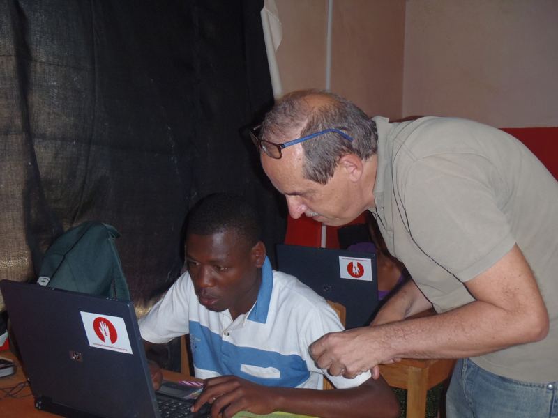 A volunteer leans over to help a young man working on a laptop in the new computer room