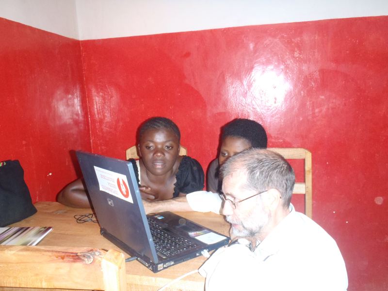 A volunteer sits beside two young women working on a laptop at a desk in the computer room