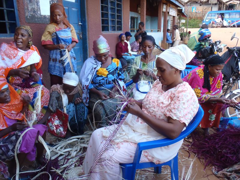 Women of the Mkeka group sit together outdoors in Bumbuli, weaving mats from dyed palm leaves