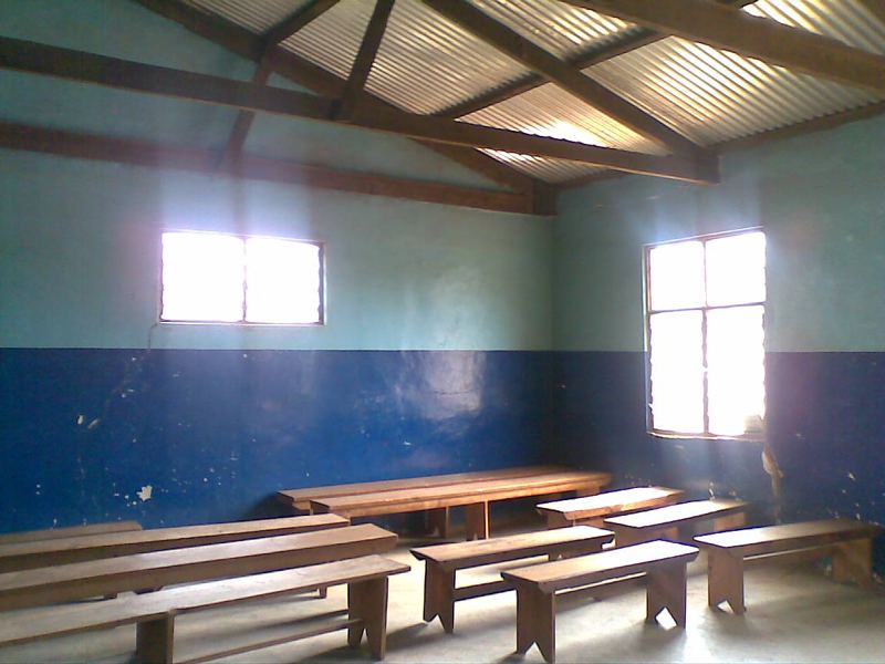 A classroom with wooden benches and desks, blue-painted walls and light from two windows